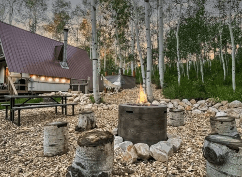 A cozy outdoor fire pit surrounded by wooden stools, with a cabin and trees in the background at dusk.